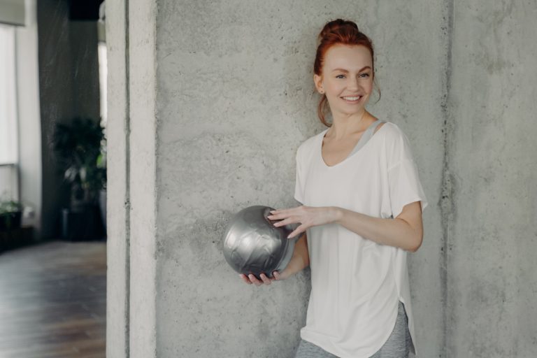 Red haired positive woman standing against gray wall with fitball in hand before pilates class