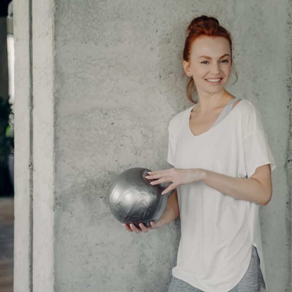 Red haired positive woman standing against gray wall with fitball in hand before pilates class